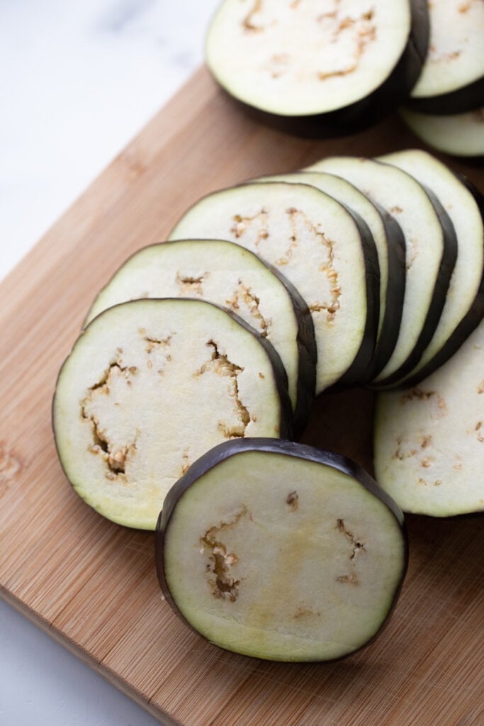 Slices of uncooked eggplant are placed on a wooden cutting board.