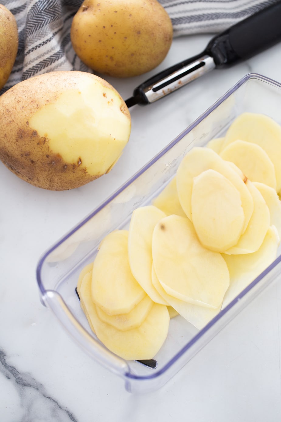 A potato is being peeled next to a glass container of sliced potatoes.
