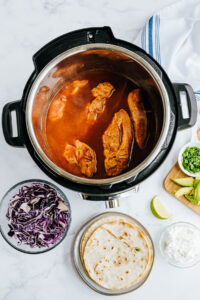 Fresh ingredients like purple cabbage and limes surround an Instant Pot containing cooked chicken.