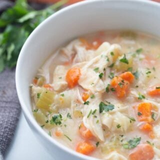 A white bowl containing chicken pot pie soup is placed on a white surface with carrots in the background.
