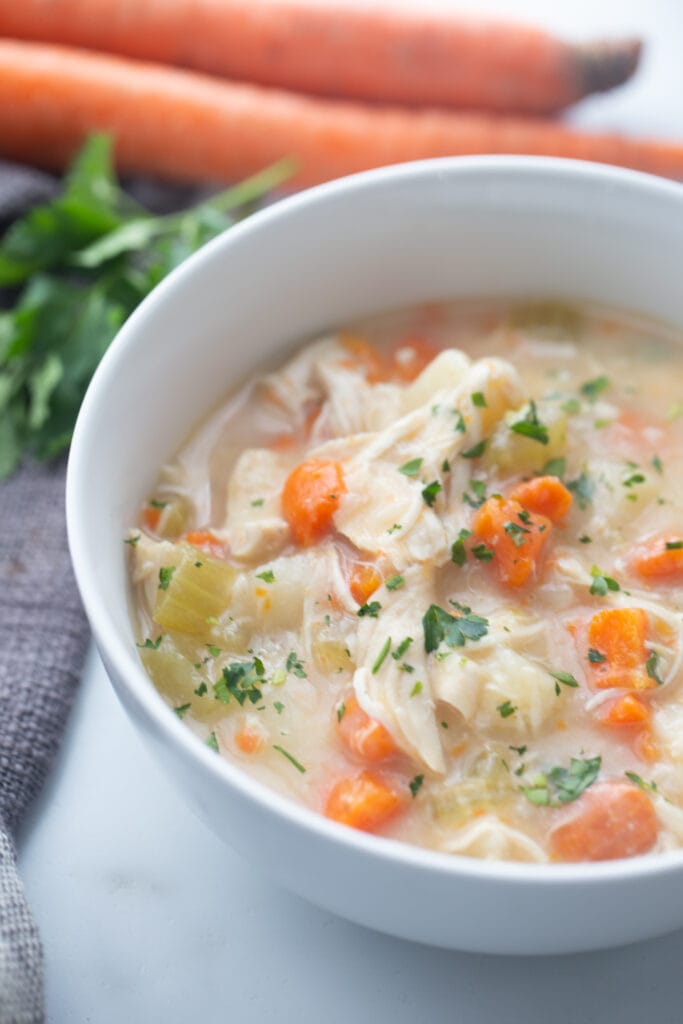 A white bowl containing chicken pot pie soup is placed on a white surface with carrots in the background. 
