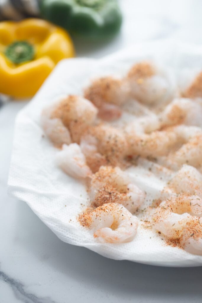Raw shrimp are being seasoned on a white paper towel.