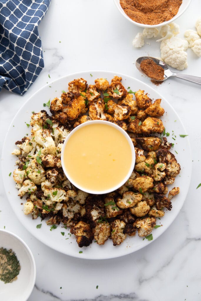 Overhead photo of seasoned cauliflower made in the air fryer. The cauliflower is on a white serving platter with a bowl of cheese sauce in the middle. 