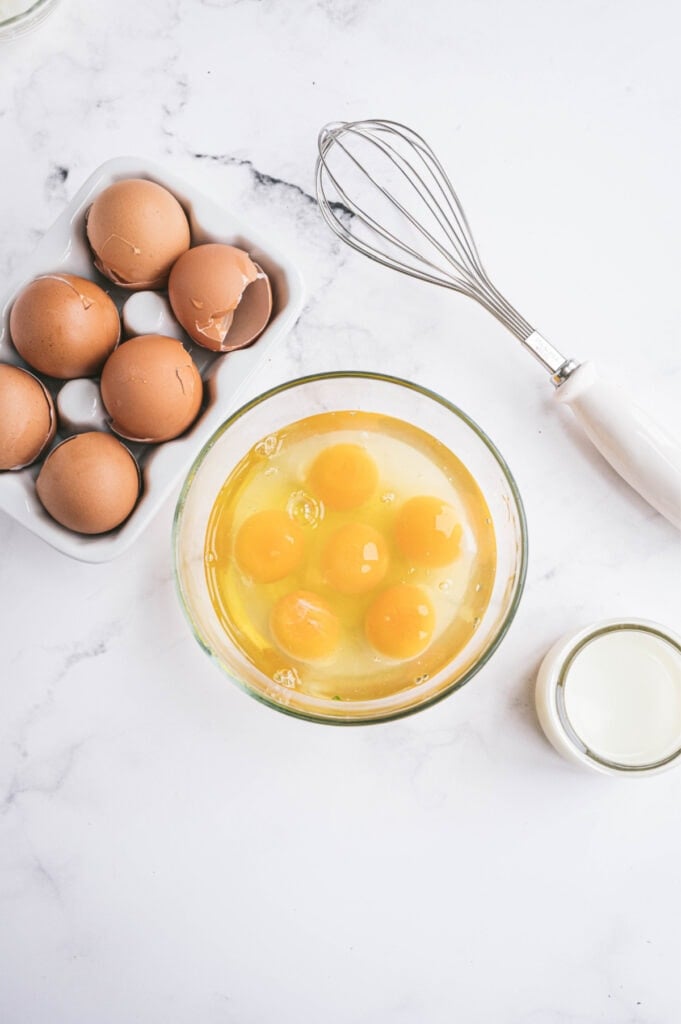 Cracked eggs are in a glass bowl next to a whisk. 