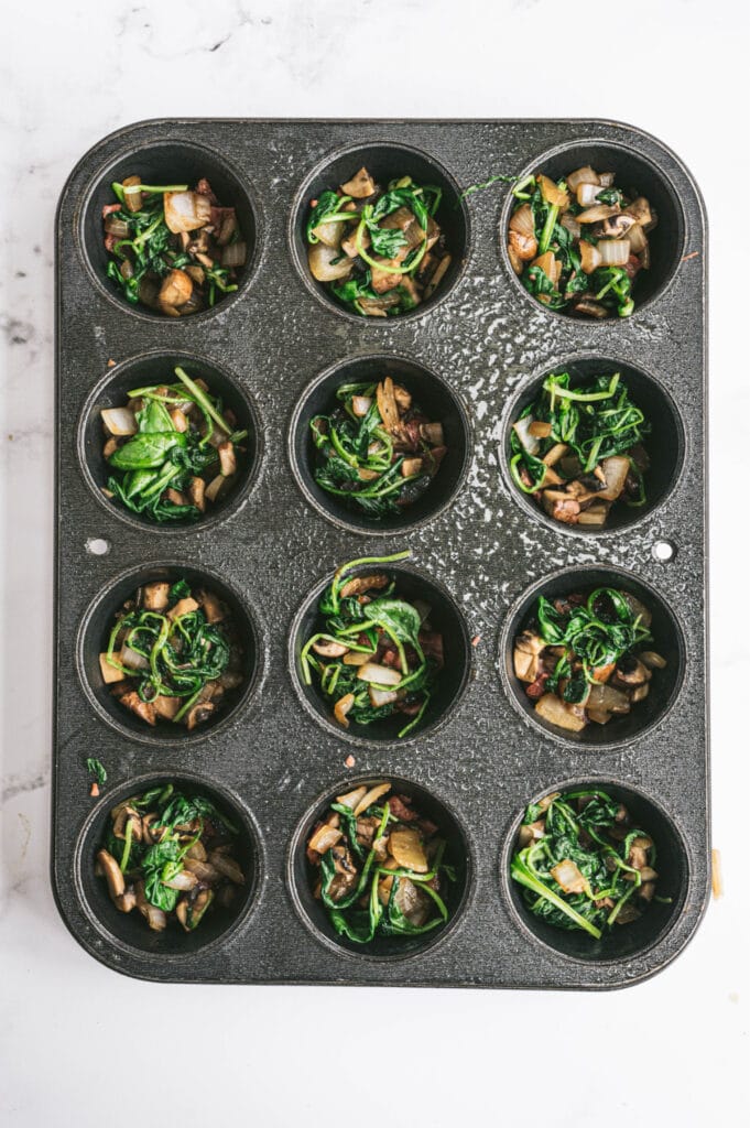 Freshly cooked veggies are placed on top of bacon bits in muffin tins.