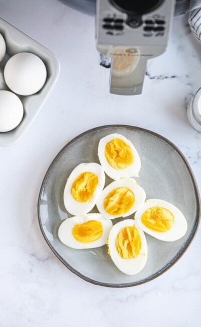 A plate of hard boiled eggs is placed next to raw eggs and an air fryer.