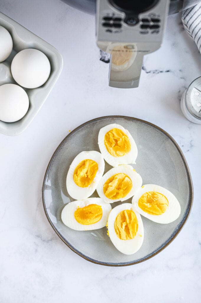 A plate of hard boiled eggs is placed next to raw eggs and an air fryer. 