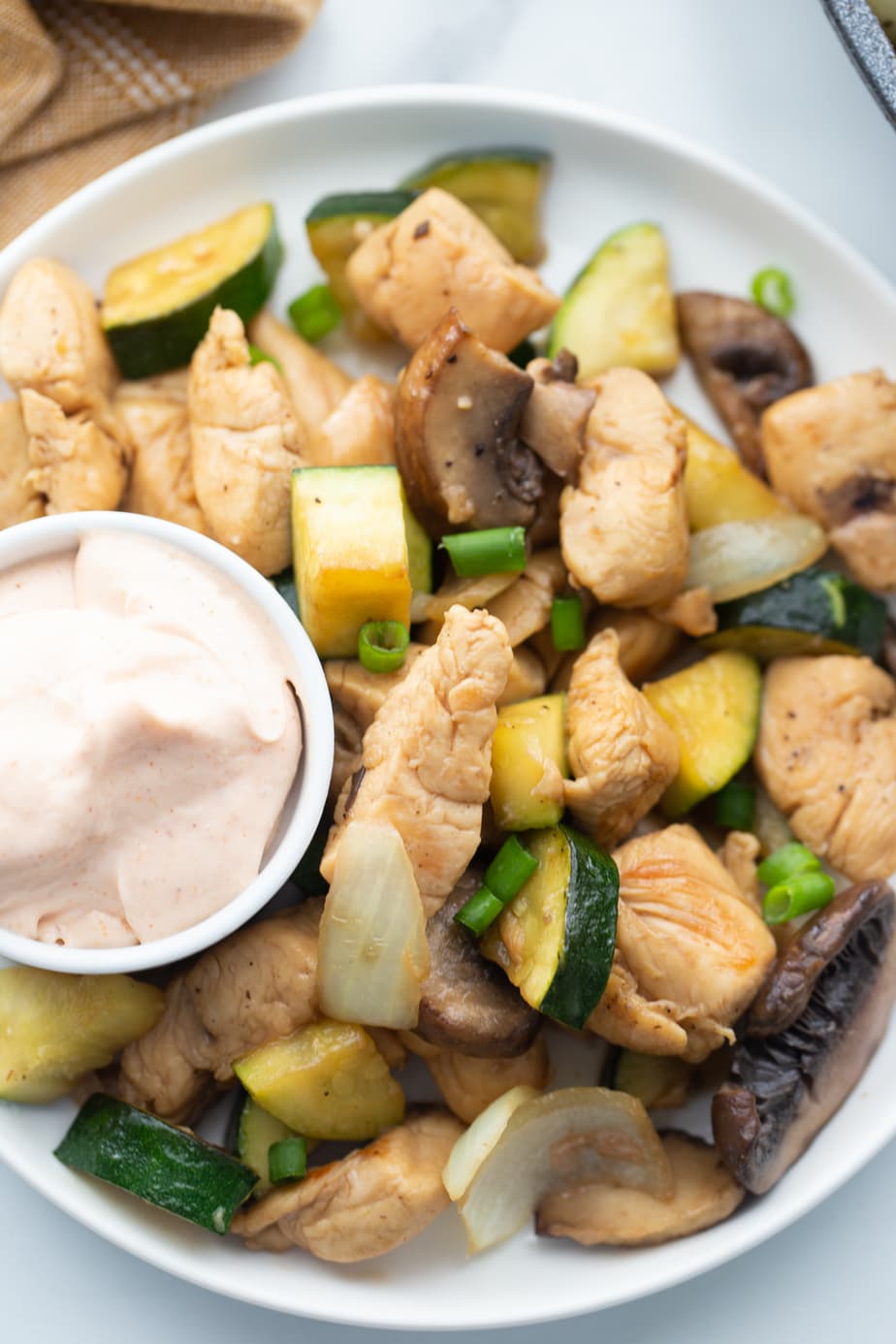 A cup of yum yum sauce sits on a white plate next to cooked chicken and vegetables.