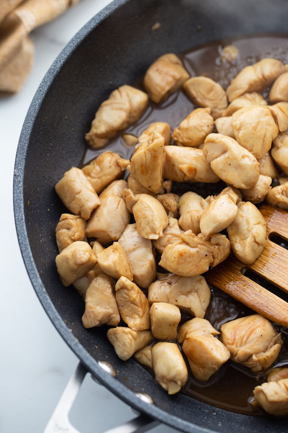 Browned chicken is being stirred by a wooden spoon in a black skillet.