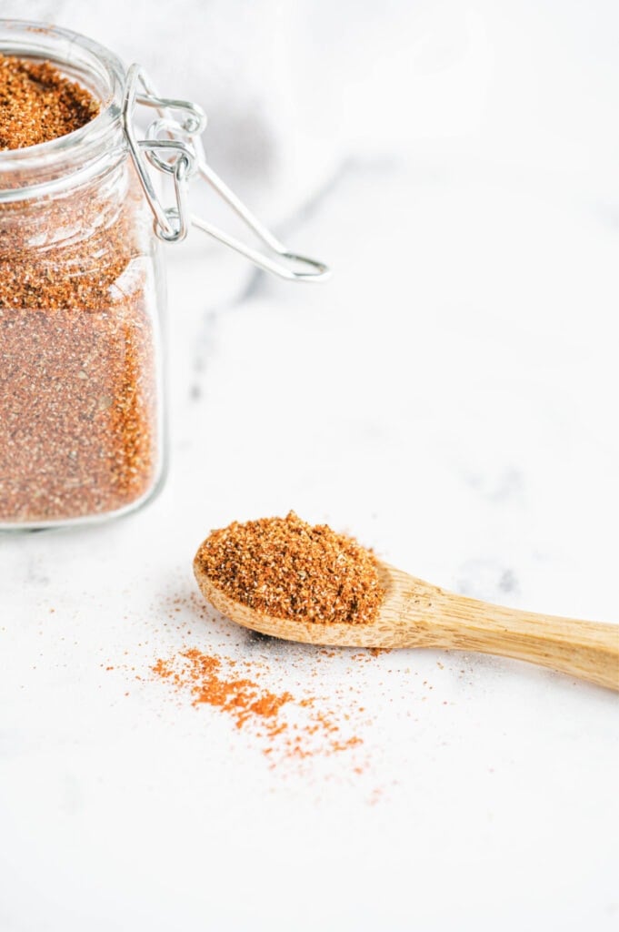 Taco seasoning blend in a glass jar on the side of the photo. A mini wooden spoon full of seasoning on a white surface.
