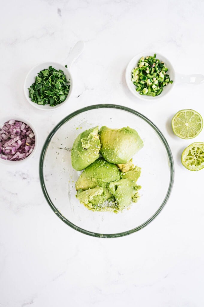 Ingredients for chipotle guacamole in glass bowls on a white marble backdrop
