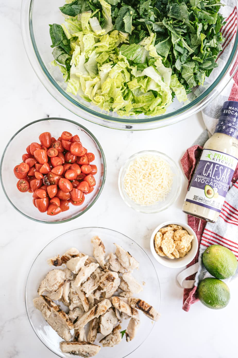 Overhead photo of ingredients in glass bowls - Greens, chopped tomatoes, diced grilled chicken, Parmesan, and Caesar dressing