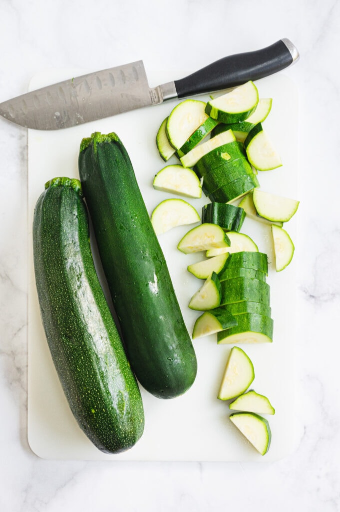 Sliced zucchini and two whole zucchini on a white cutting board with a chef knife laying above them 