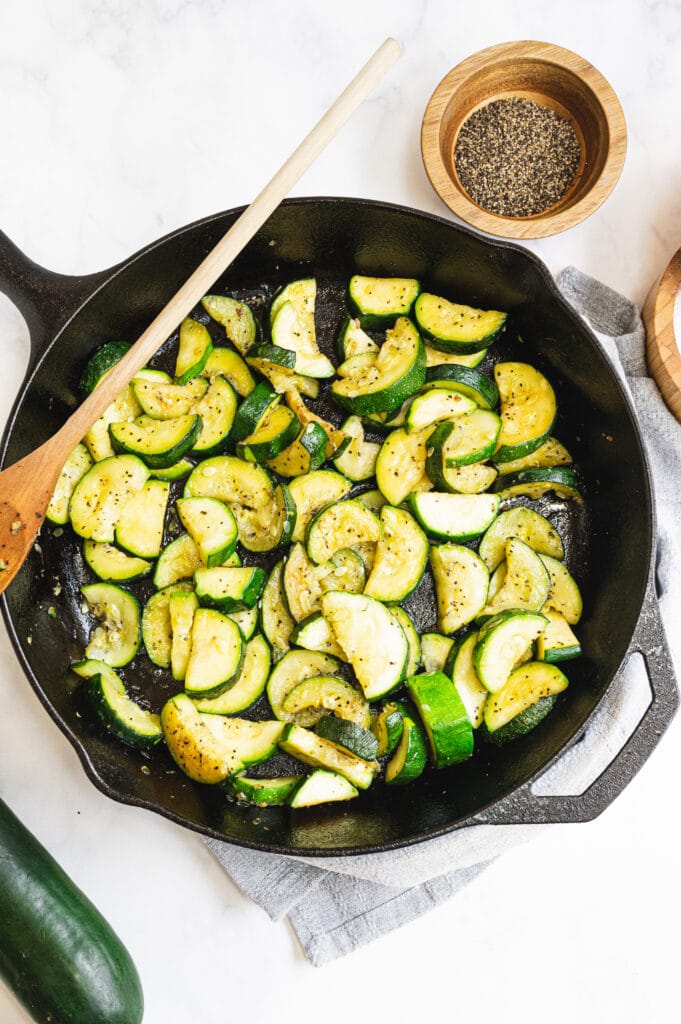 Overhead shot of sauteed zucchini in a cast iron pan with a wooden spoon resting on the side