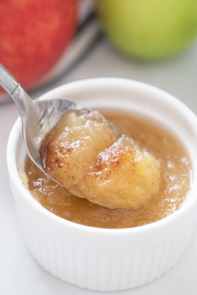 Close up of a spoon of applesauce and a white ramekin with apples in the background 