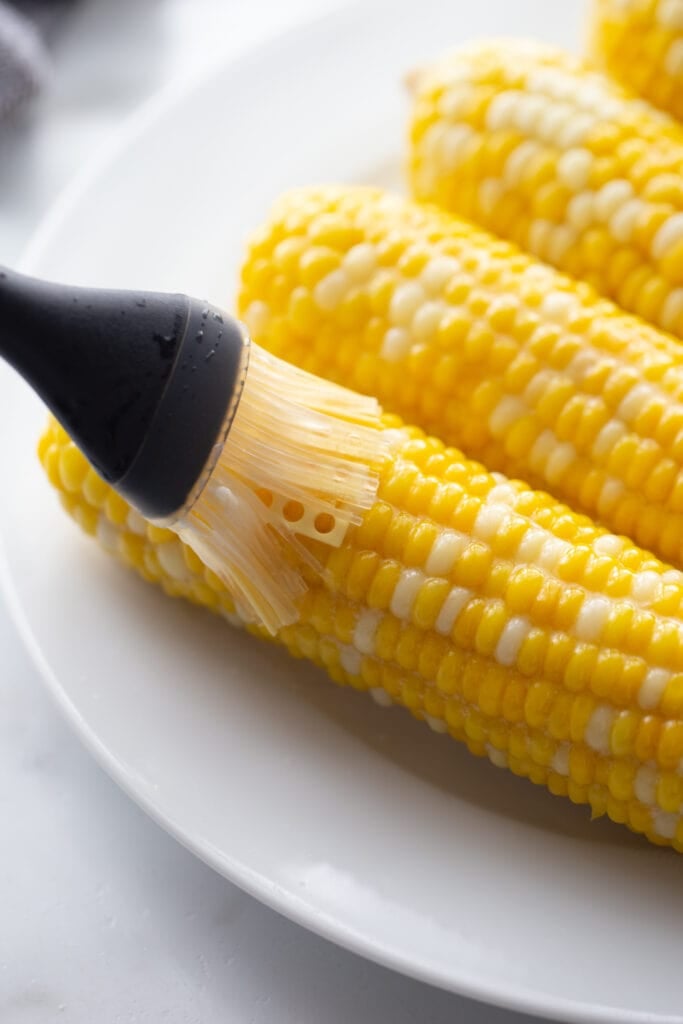 Cooked corn on the cob on white plate, being brushed with butter