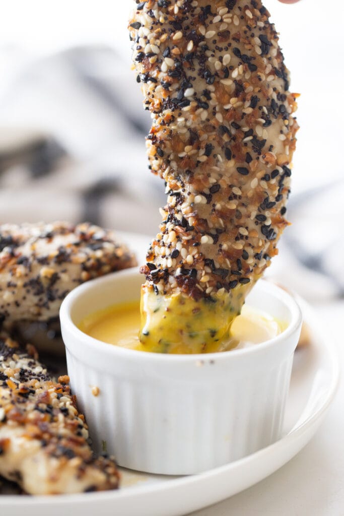 A close up of a chicken tender covered in everything bagel seasoning being dipped into maple mustard sauce. 