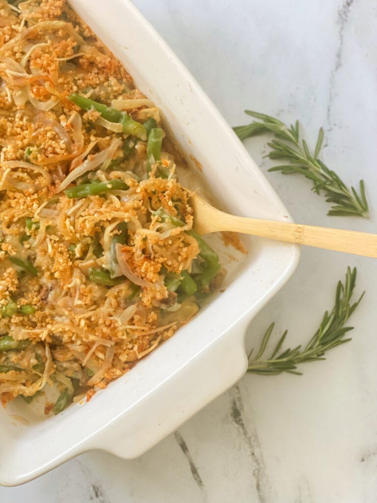 Overhead image: green bean casserole with crispy onion topping in a white casserole dish with a wooden spoon.