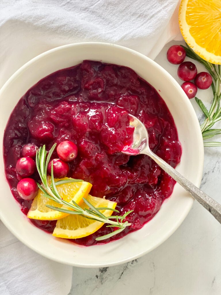 Overhead image: Homemade cranberry sauce in a white bowl, garnished with orange slices, whole cranberries, and sprigs of rosemary.