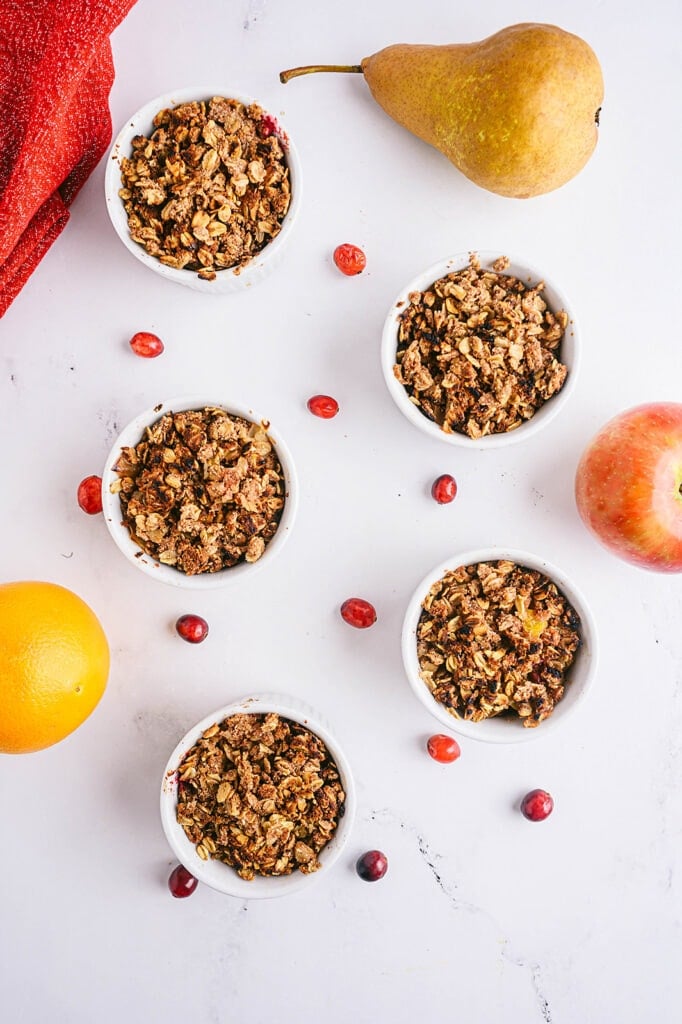 Overhead shot of four white ramekins with pear apple cranberry crisp topped with oats and with fresh cranberries and apples scattered around the table.
