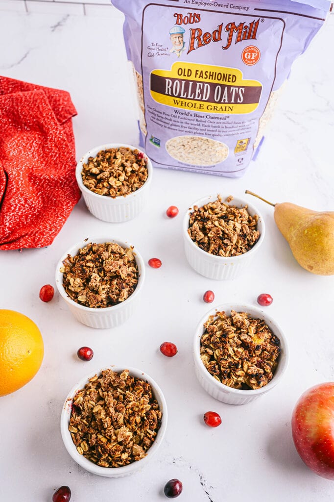 Overhead shot of four white ramekins with pear apple cranberry crisp topped with coconut whipped cream and with fresh cranberries and apples scattered around the table. A bag of Bob's Red Mill Old Fashioned Oats sits in the background.