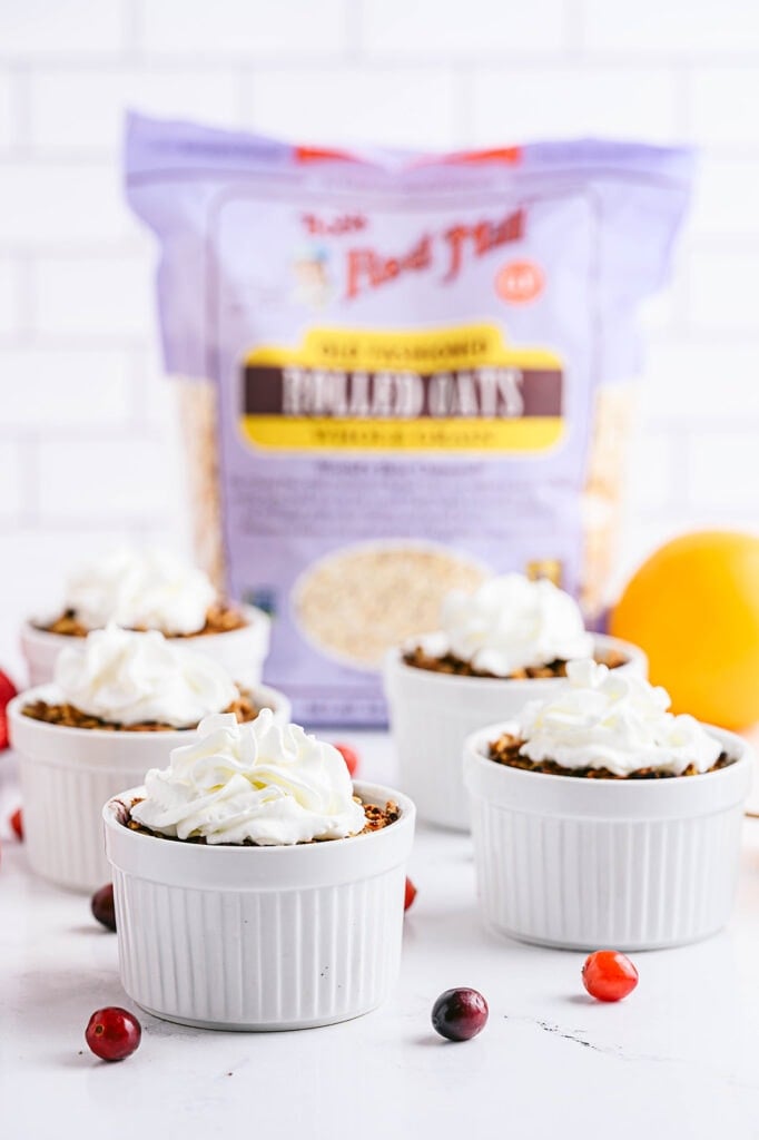 Close up of four white ramekins with pear apple cranberry crisp topped with coconut whipped cream and with fresh cranberries and apples scattered around the table.  A bag of Bob's Red Mill Old Fashioned Oats sits in the background.