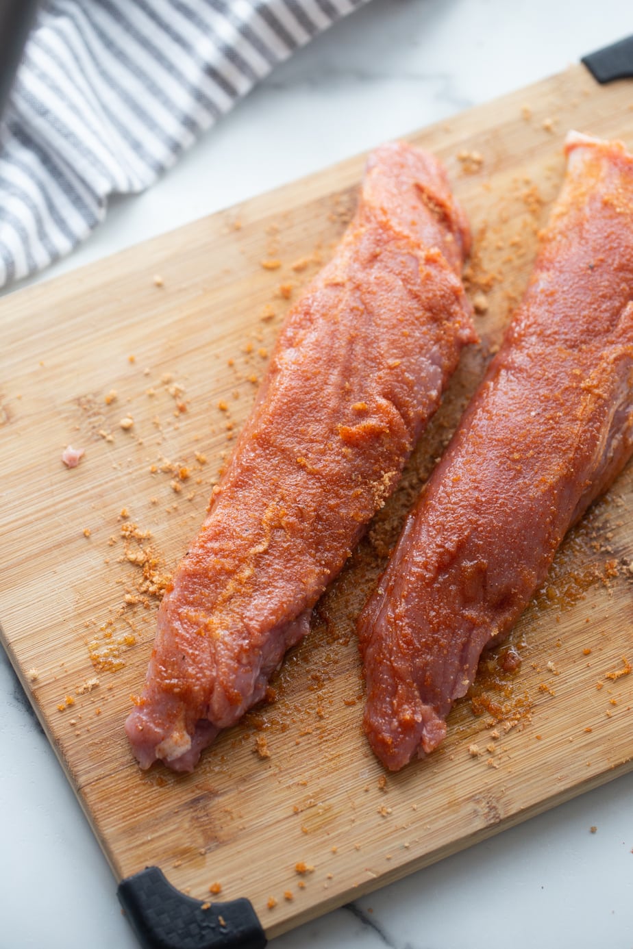 Tow raw pork tenderloins on a wooden cutting board coated with spice rub. 