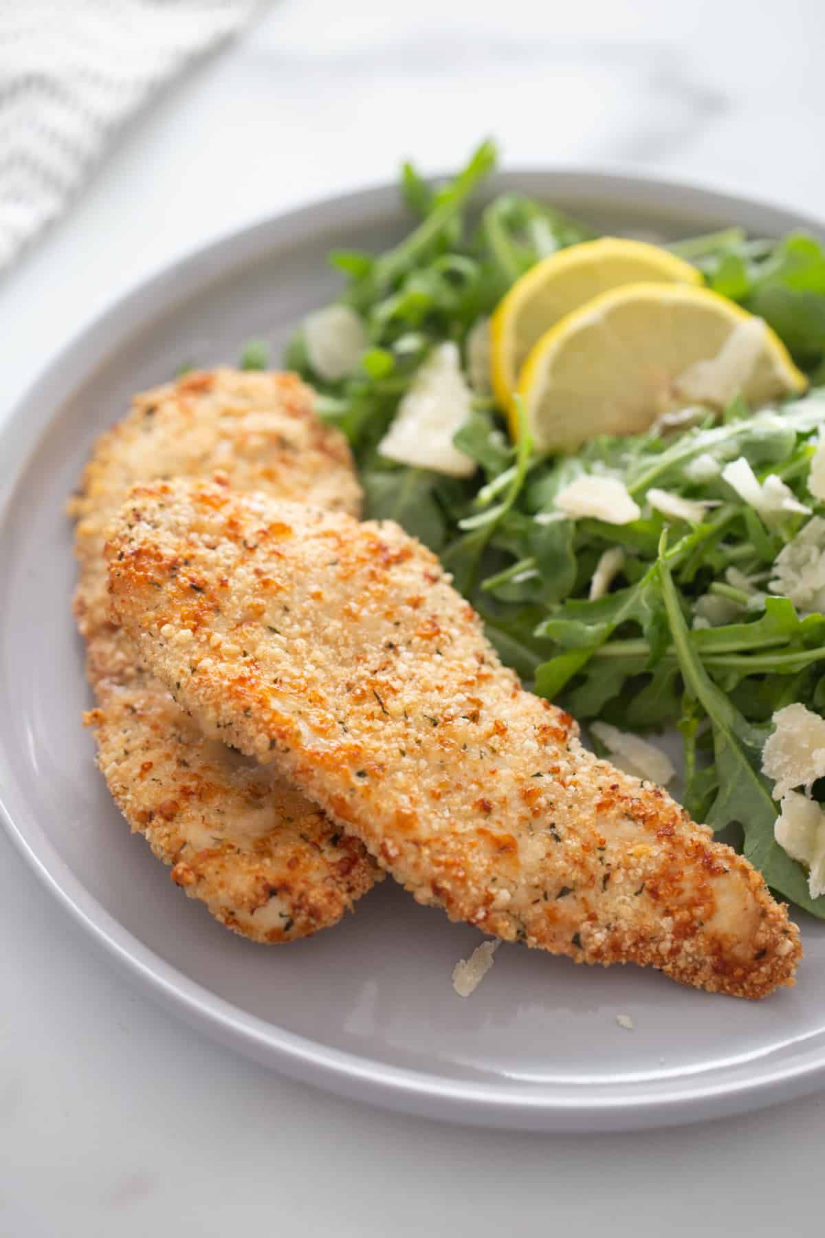 Two Chicken cutlets on a white pate served with arugula salad 