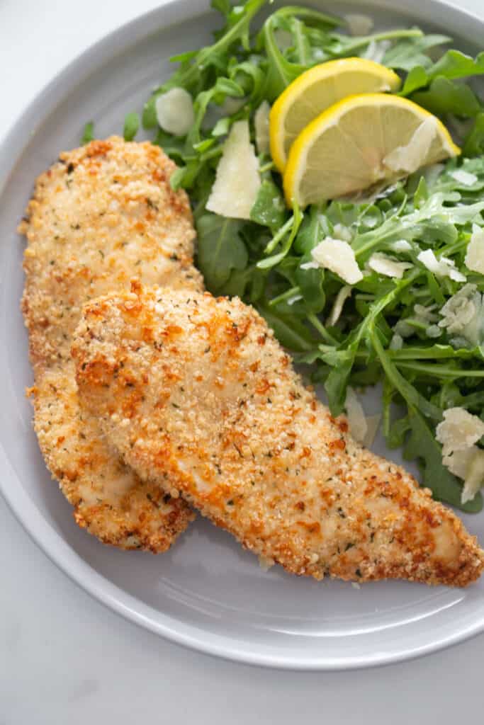Plate of Chicken Cutlets next to an arugula salad topped with shredded parm and lemon 