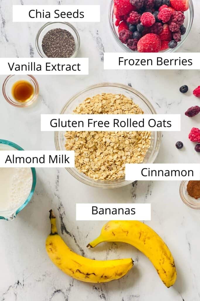 Flatlay of ingredients for One Pan Baked Oatmeal laying on a table with text overtop of each item:  bananas, gluten free oats, vanilla extract, frozen berries, almond milk, cinnamon. 
