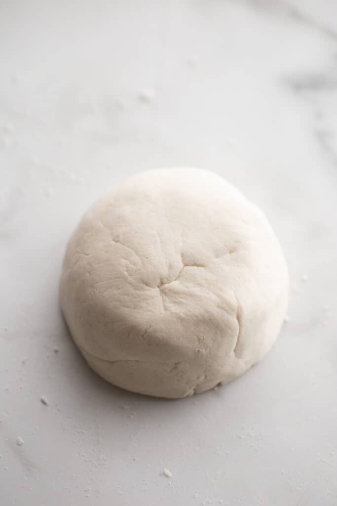 Pizza dough that has been kneaded and smoothed out is on a white counter top. 