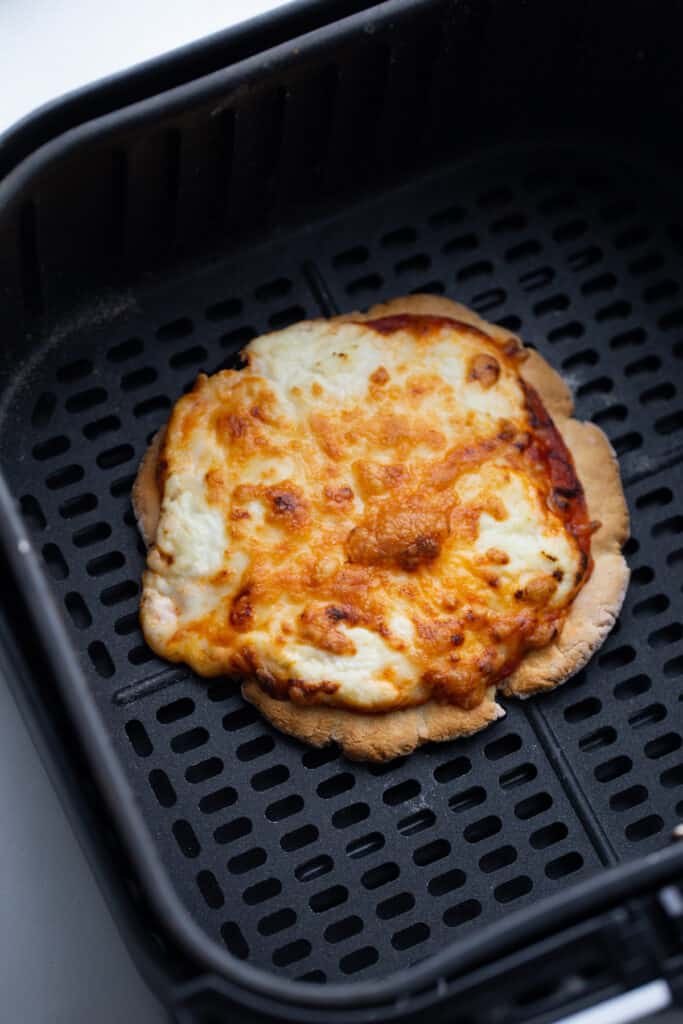 A small personal pizza with melted crispy cheese sits in an air fryer basket. 