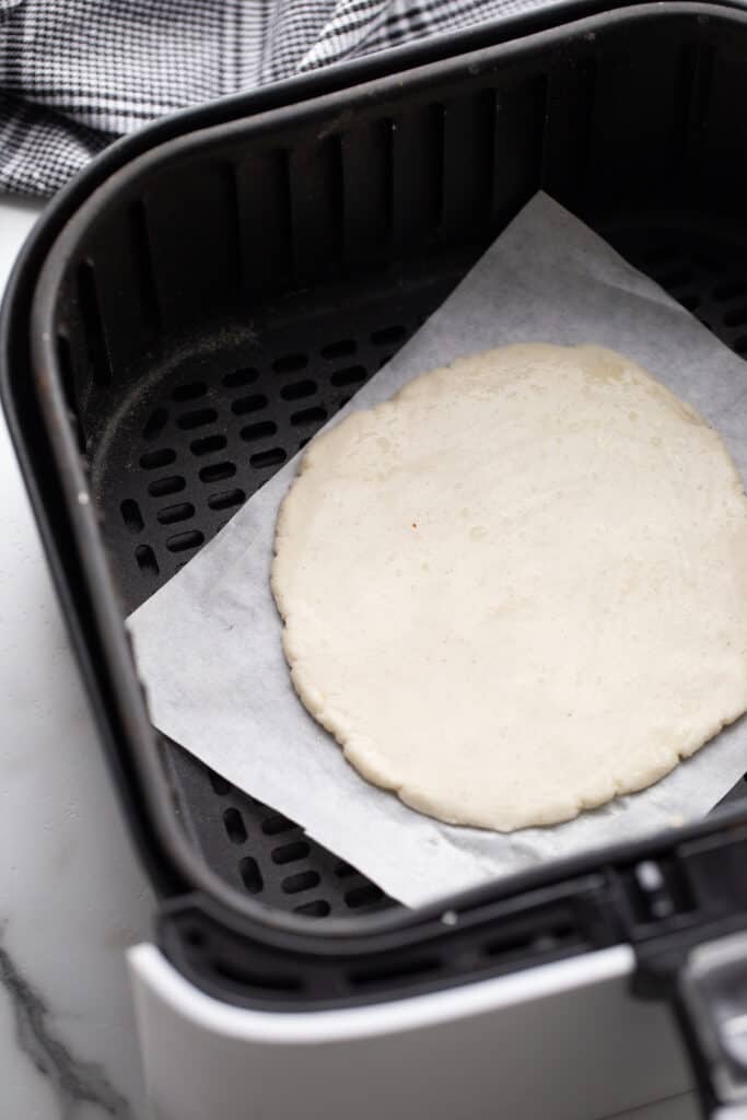 Pizza crust in an air fryer basket on top of parchment paper. 