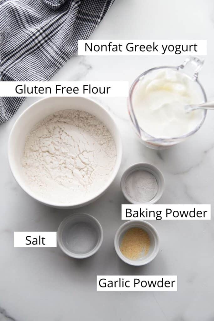 Ingredients for gluten free greek yogurt pizza crust sit on a table: gluten free flour, greek yogurt, baking powder, salt, and garlic powder. 