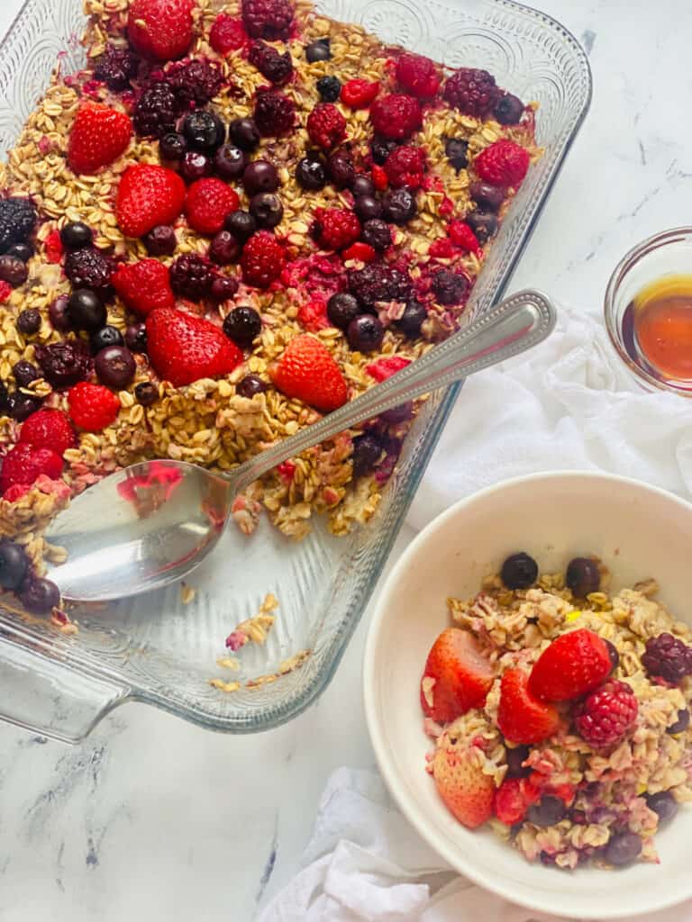 One pan of baked oats topped with berries next to a bowl of one portion of the oats on a marble counter.  
