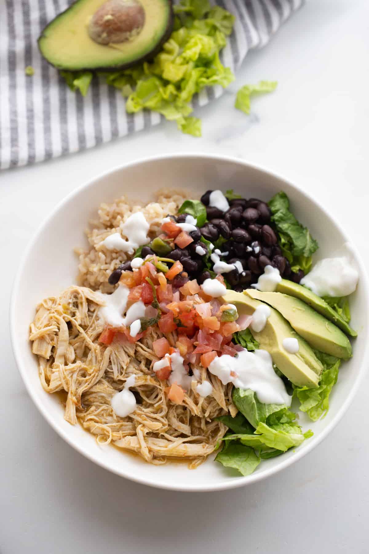 A bowl of salsa verde chicken topped with Greek yogurt, brown rice, pico de gallo, greens, avocado on a white table with a blue striped napkin nearby. 