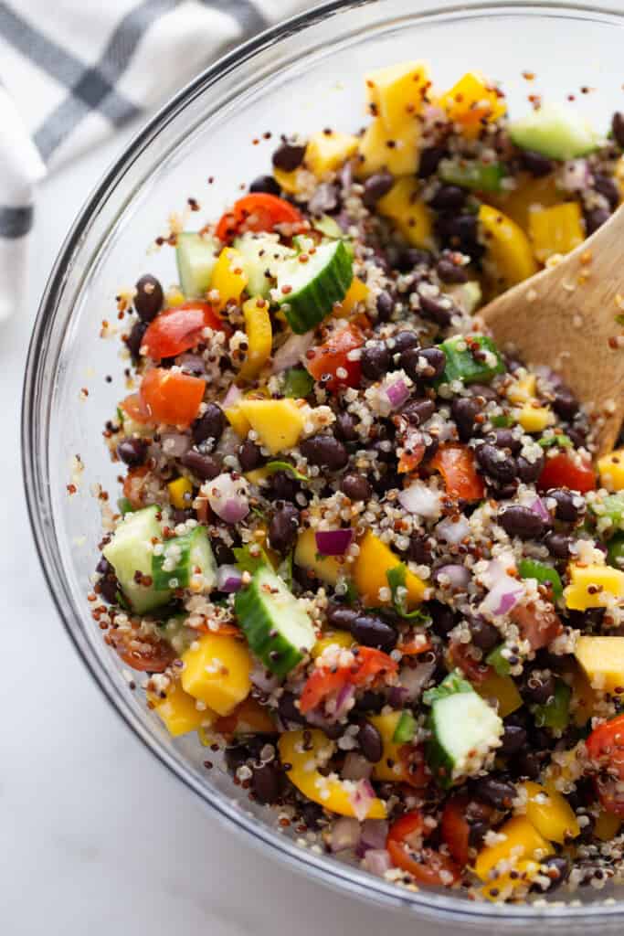 Cooked quinoa, black beans, cucumbers, tomatoes, mangoes, and onions all mixed in a colorful salad in a clear bowl.