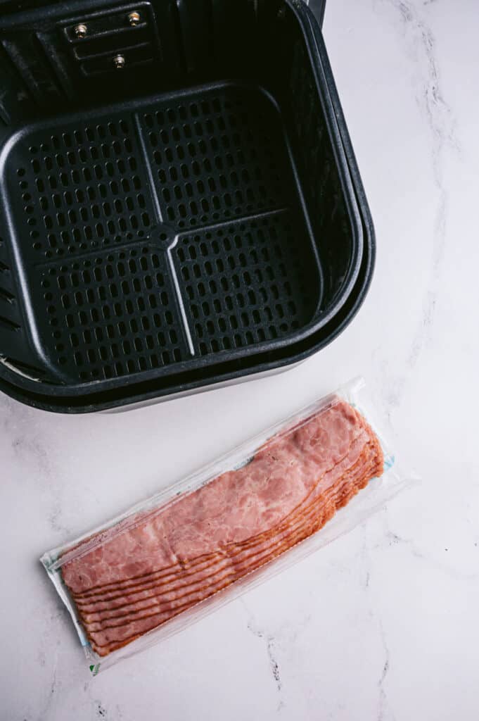 An empty air fryer basket and a package of Turkey Bacon sitting on a marble table. 