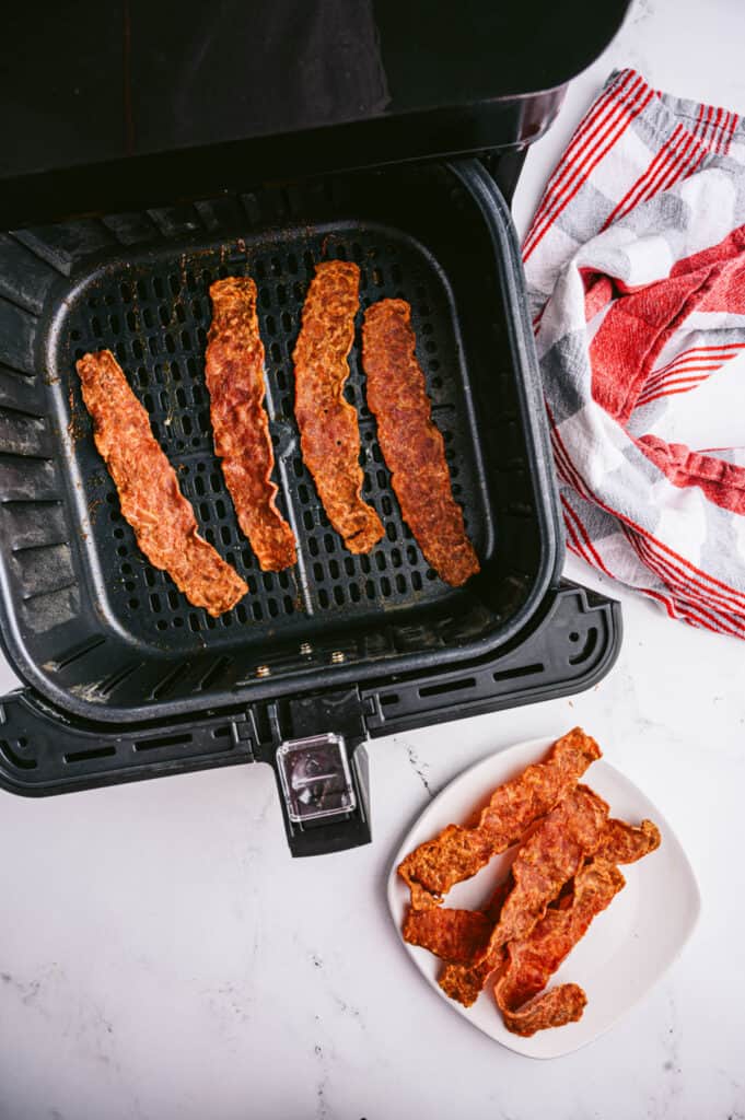 Four strips of turkey bacon cooked to crispy perfection in an air fryer basket with four more pieces sitting on a white plate nearby. 