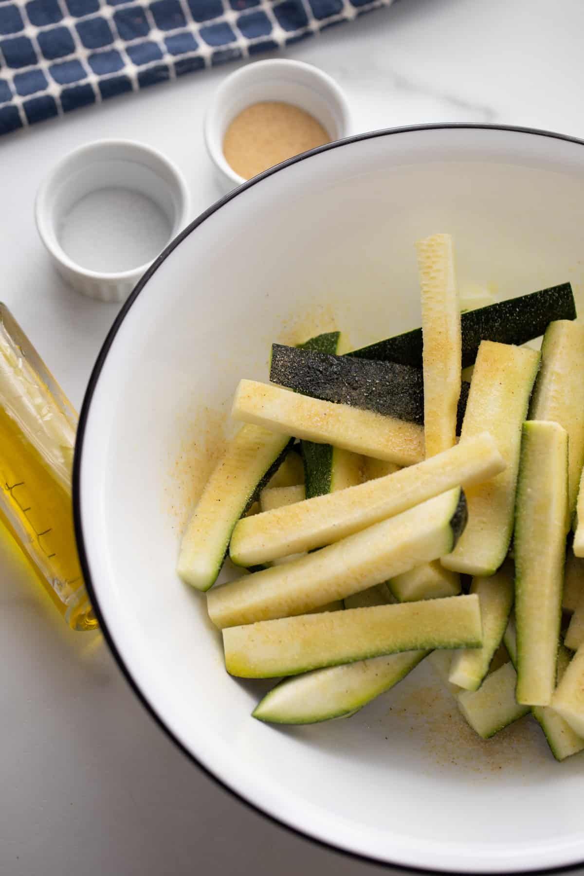 A shallow white bowl with zucchini cut into sticks with salt and garlic powder sitting to the side