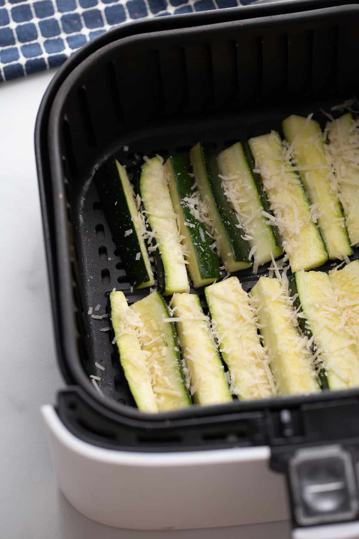 Sliced zucchini arranged in one layer inside of an air fryer basket and topped with shredded parmesan cheese