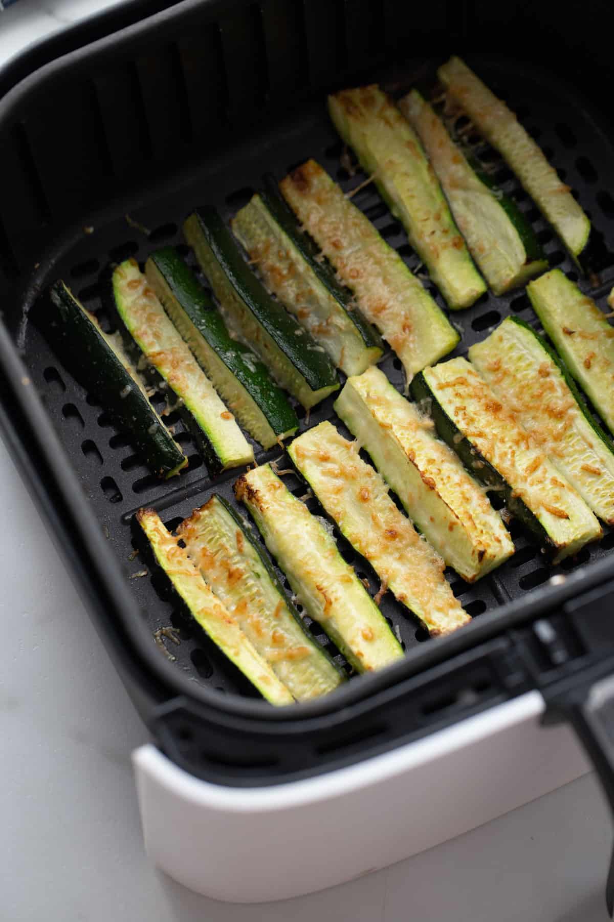 Two rows of cooked zucchini cut into french fry shapes inside of an air fryer