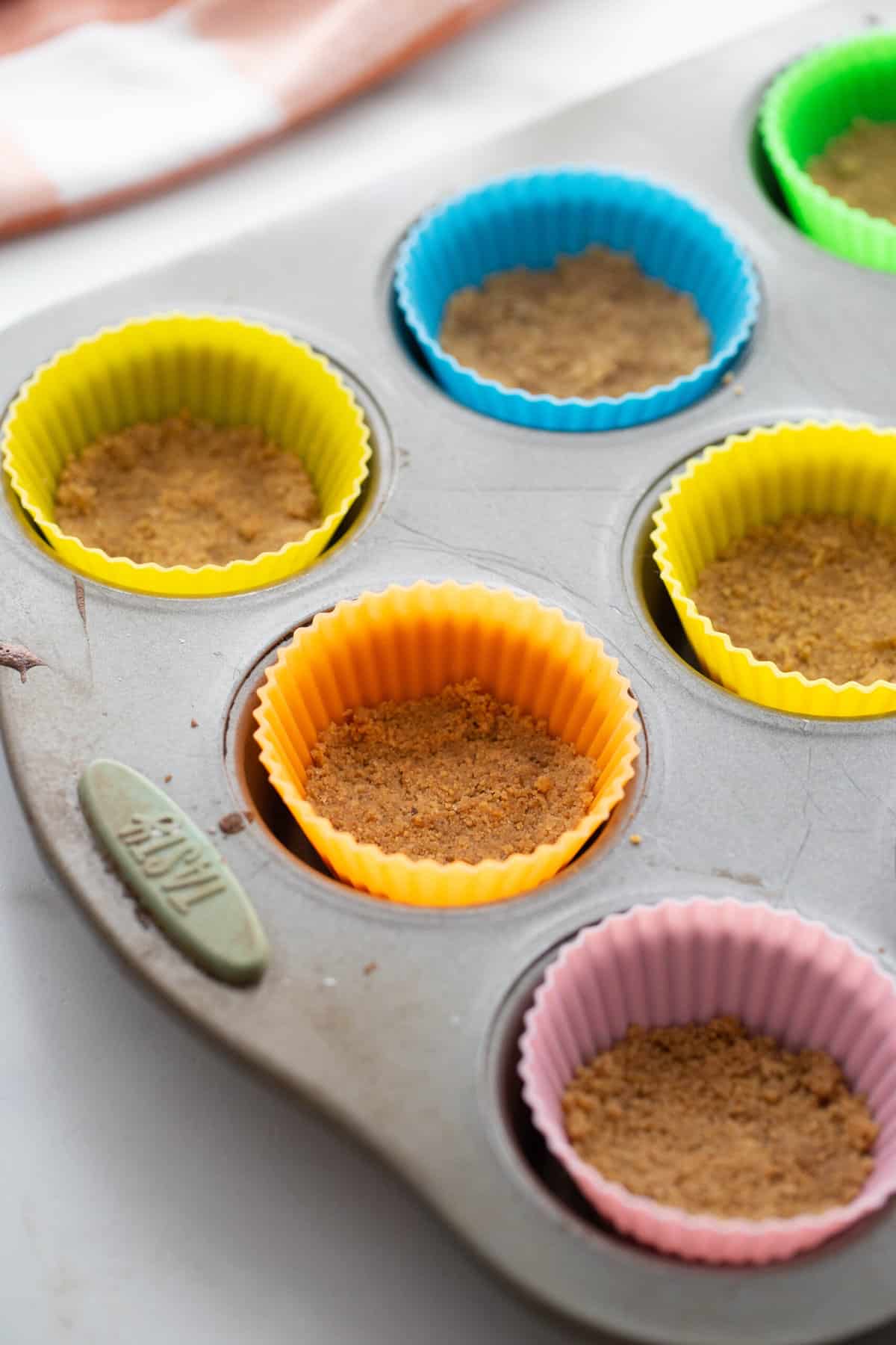 A gluten free graham cracker crust inside of colorful silicone cupcake liners. 