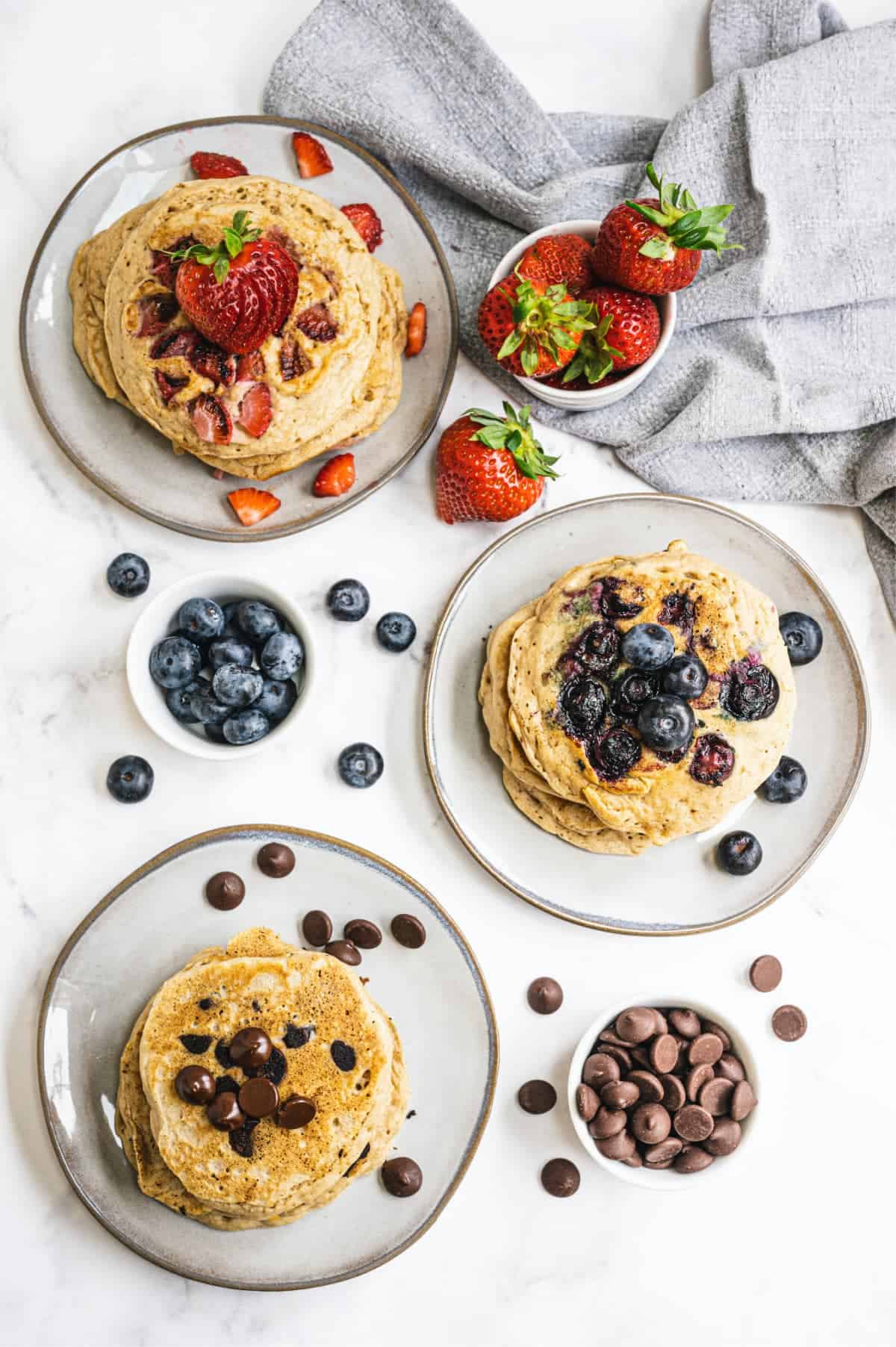Three plates with short stacks of pancakes, each with a different topping/mix in - Strawberries, blueberries, and chocolate chips.