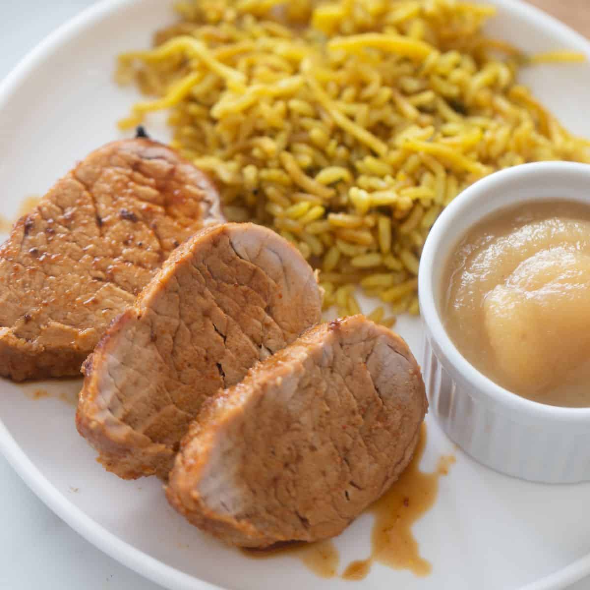 Plate with sliced air fryer pork tenderloin drizzled with juice, a side of homemade applesauce, and rice pilaf.