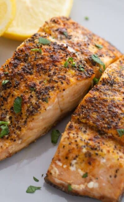 A white plate with seasoned air fryer salmon garnished with two slices of lemon on the side.
