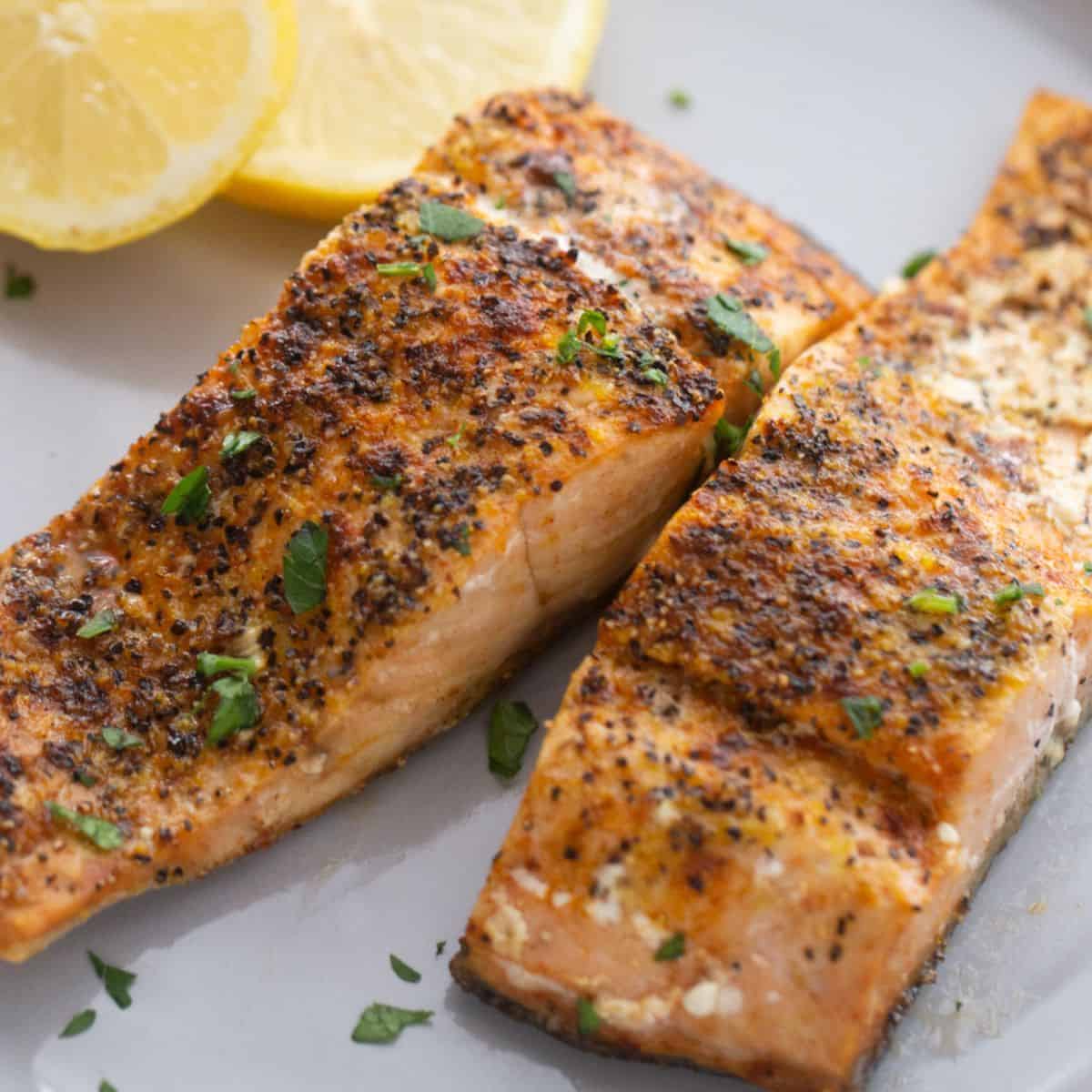 A plate of two air fryer salmon fillets seasoned with lemon pepper seasoning and garnished with fresh herbs and lemon. 