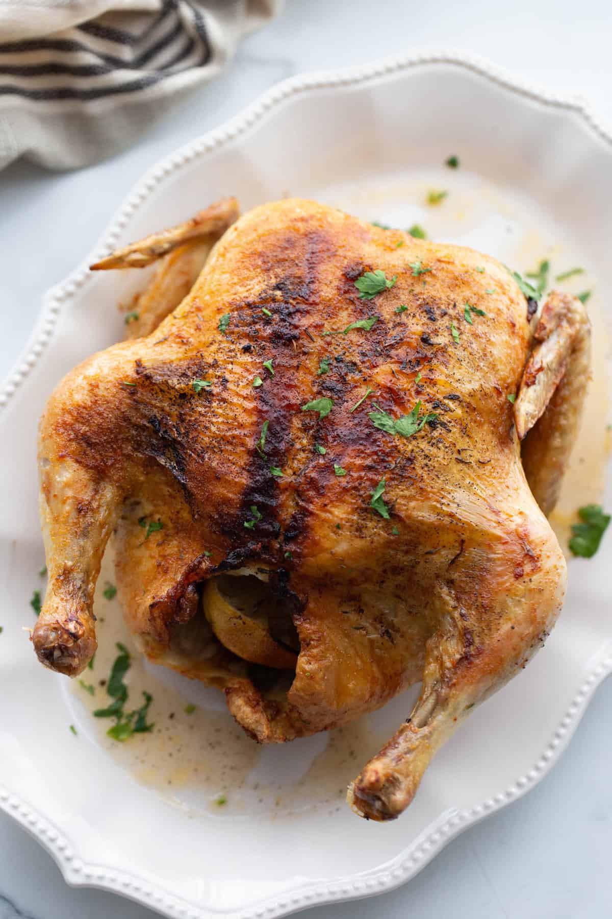 A cooked whole chicken on a white plate garnished with fresh herbs.