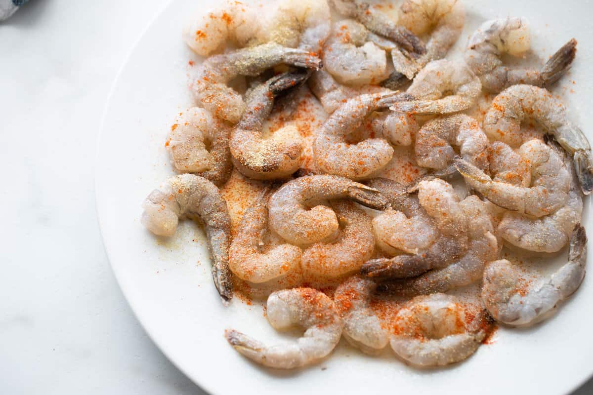 Raw shrimp on a white plate seasoned with paprika, garlic powder, and salt.