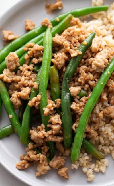A plate of ground chicken stir fry on a bed of brown rice and stopped with steamed green beans.